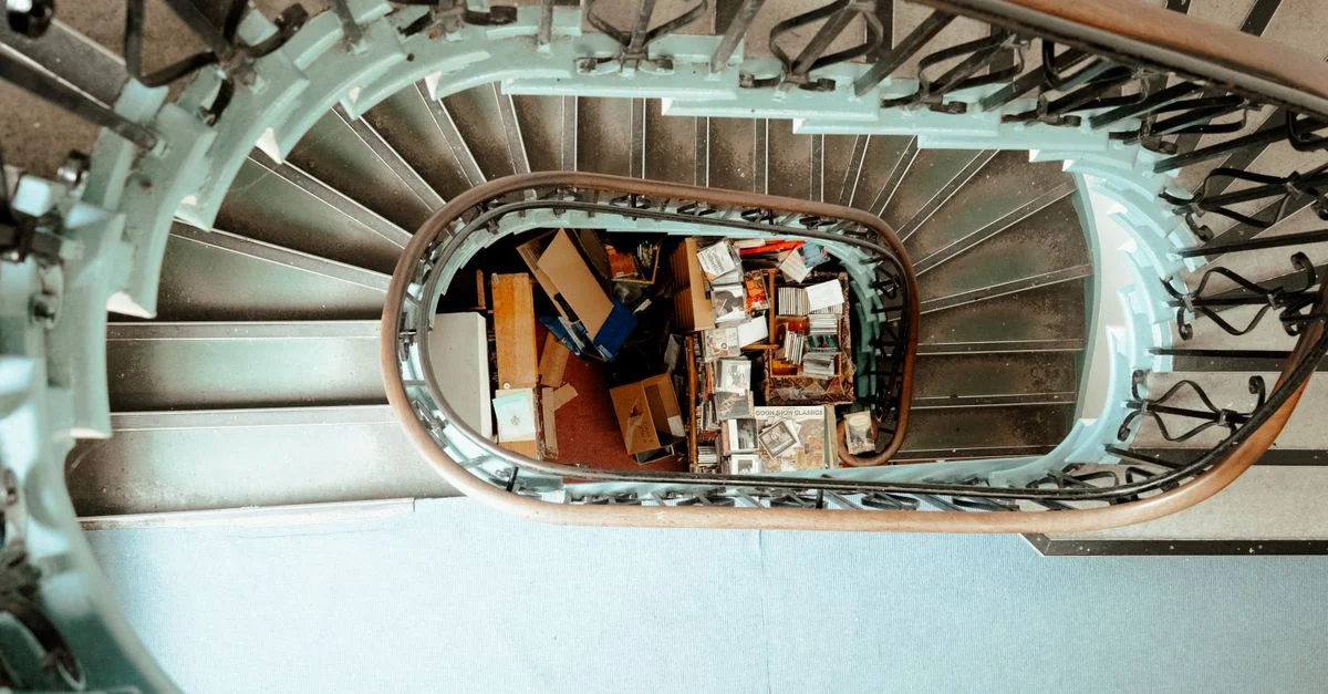 Vue de dessus d'un escalier en colimaçon avec des boîtes et des objets en désordre au centre, créant une géométrie dynamique.