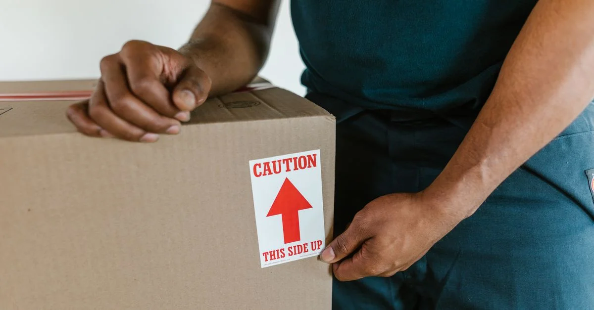 Une personne manipulant une boîte de déménagement avec une étiquette 'précaution, ce côté vers le haut' à l'intérieur.