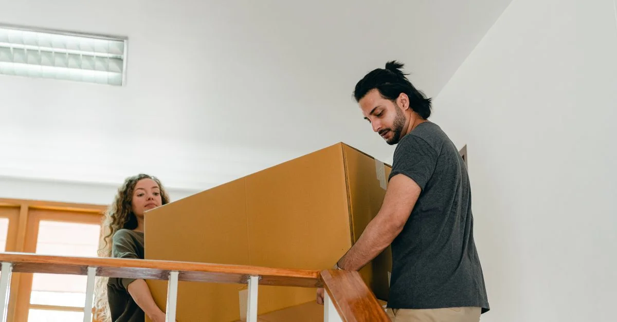 Un couple portant une grande boîte en carton dans les escaliers, symbolisant un déménagement dans une nouvelle maison.